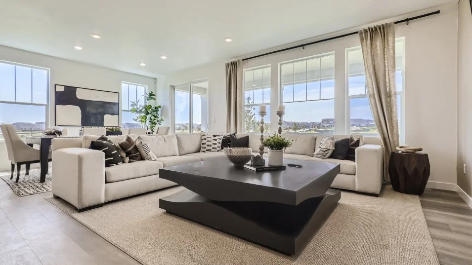 This is a bright and spacious living room featuring a large sectional sofa with decorative pillows, a unique geometric coffee table, and neutral-toned decor. Large windows with sheer curtains provide ample natural light and views of the outside. The room is carpeted and has a modern, inviting feel, perfect for relaxation and entertaining.