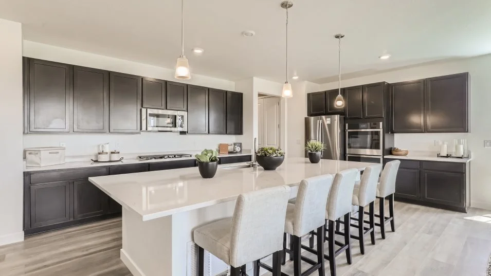 This is a modern kitchen with dark cabinetry and stainless steel appliances. A large island with a white countertop provides ample space for food preparation and seating with four bar stools. Pendant lights hang above the island, adding a touch of elegance to the space.