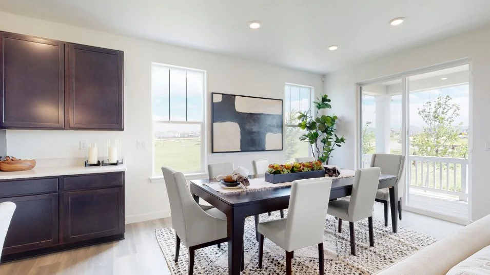 This is a bright and airy dining room featuring a dark wood dining table with light gray upholstered chairs. A modern abstract painting hangs on the wall, complemented by a large plant in the corner. The room is well-lit with natural light streaming through the windows and sliding glass doors, which lead to an outdoor balcony.