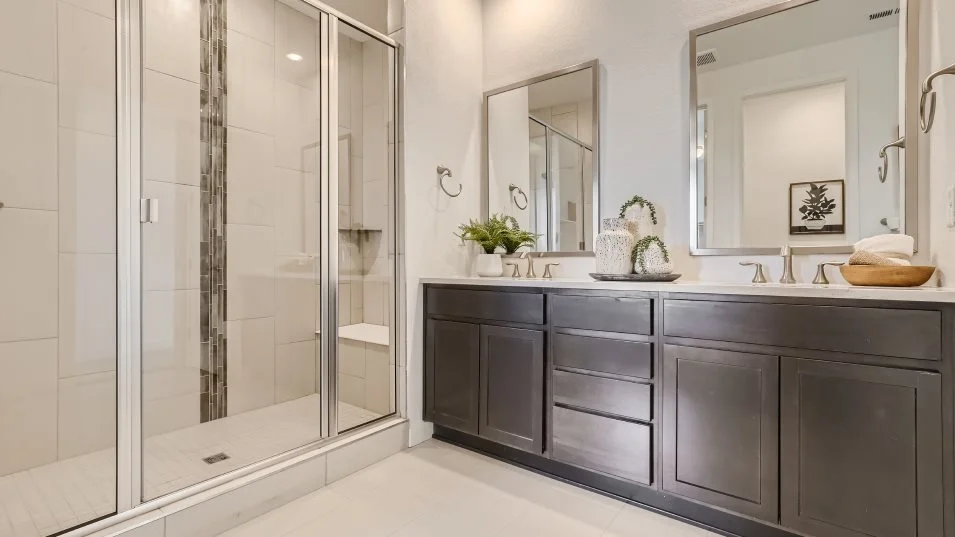 This is a primary bathroom featuring a glass-enclosed shower with white and gray tile, a double vanity with dark gray cabinets and white countertops, and two framed mirrors. The bathroom has a clean and modern aesthetic, with neutral colors and minimalist decor. The space appears bright and well-maintained, suggesting a luxurious and comfortable experience.