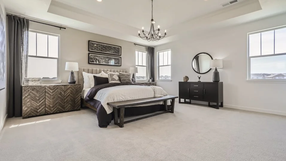 This is a spacious primary bedroom featuring a neutral color palette with a large bed as the focal point. The room is well-lit with natural light from multiple windows and features a chandelier, adding a touch of elegance. The furniture includes a dresser, a bench at the foot of the bed, and decorative wall art, creating a comfortable and stylish retreat.