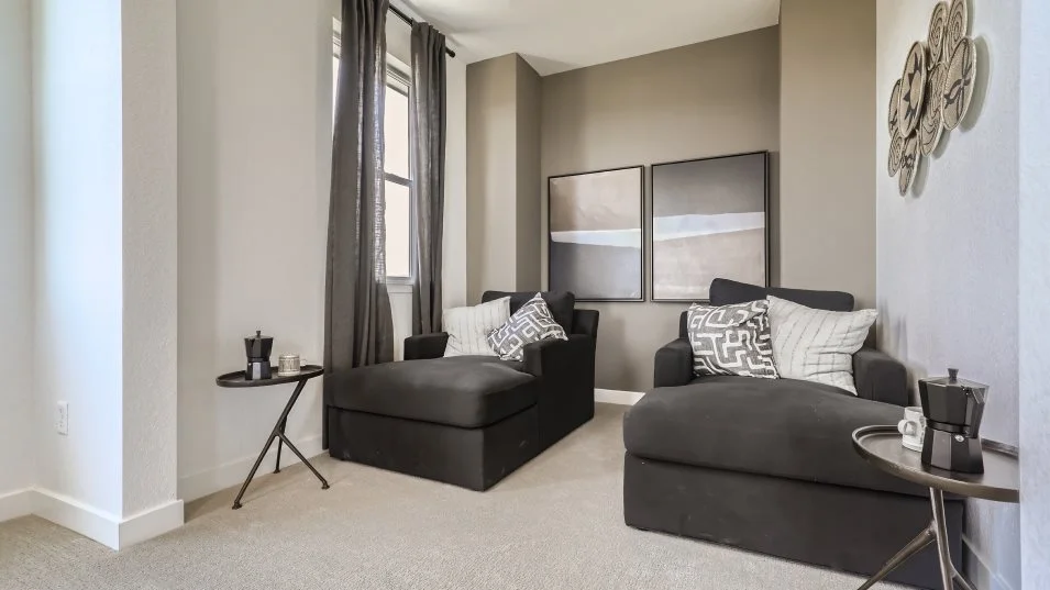 This interior shot showcases a cozy living room setup with two dark-colored chaise lounges, each adorned with decorative pillows. A small side table with a coffee maker sits beside each lounge, adding a touch of convenience. The room is decorated with framed artwork and wall decor, creating a stylish and inviting atmosphere.
