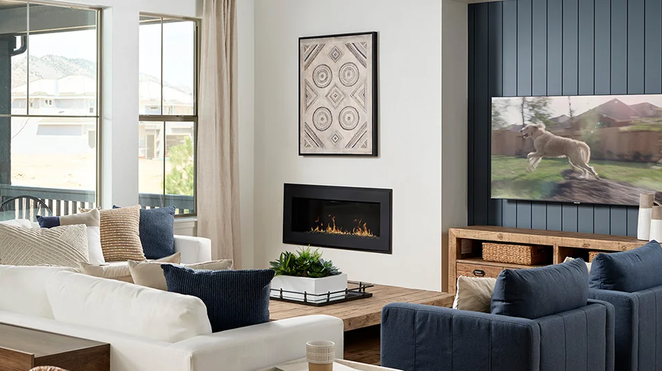This is a cozy living room featuring a modern fireplace with a geometric art piece above it. The room is furnished with a white sofa, blue armchairs, and a wooden coffee table, creating a comfortable and inviting atmosphere. A large television is mounted on a blue accent wall, and natural light streams in through the windows.