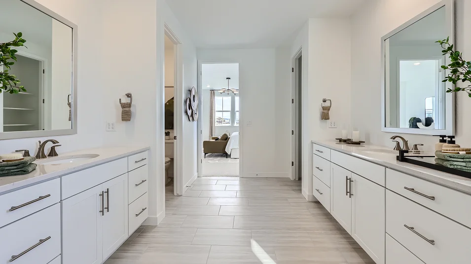 This is a bright and spacious primary bathroom featuring dual vanities with white cabinetry and light countertops. The bathroom has a clean, modern aesthetic with tile flooring and neutral color palette. A hallway leads to the bedroom, creating a seamless transition between spaces.
