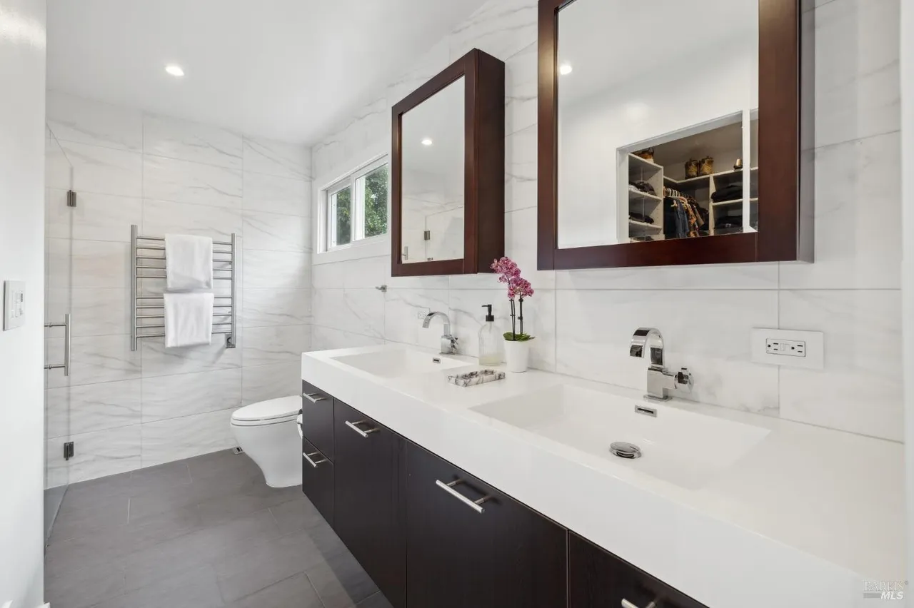 This is a well-lit primary bathroom featuring a double vanity with a white countertop and dark wood cabinetry. Two framed mirrors hang above the sinks, and the walls are covered in white marble-like tiles. A heated towel rack is visible to the left, and the overall impression is clean and modern.