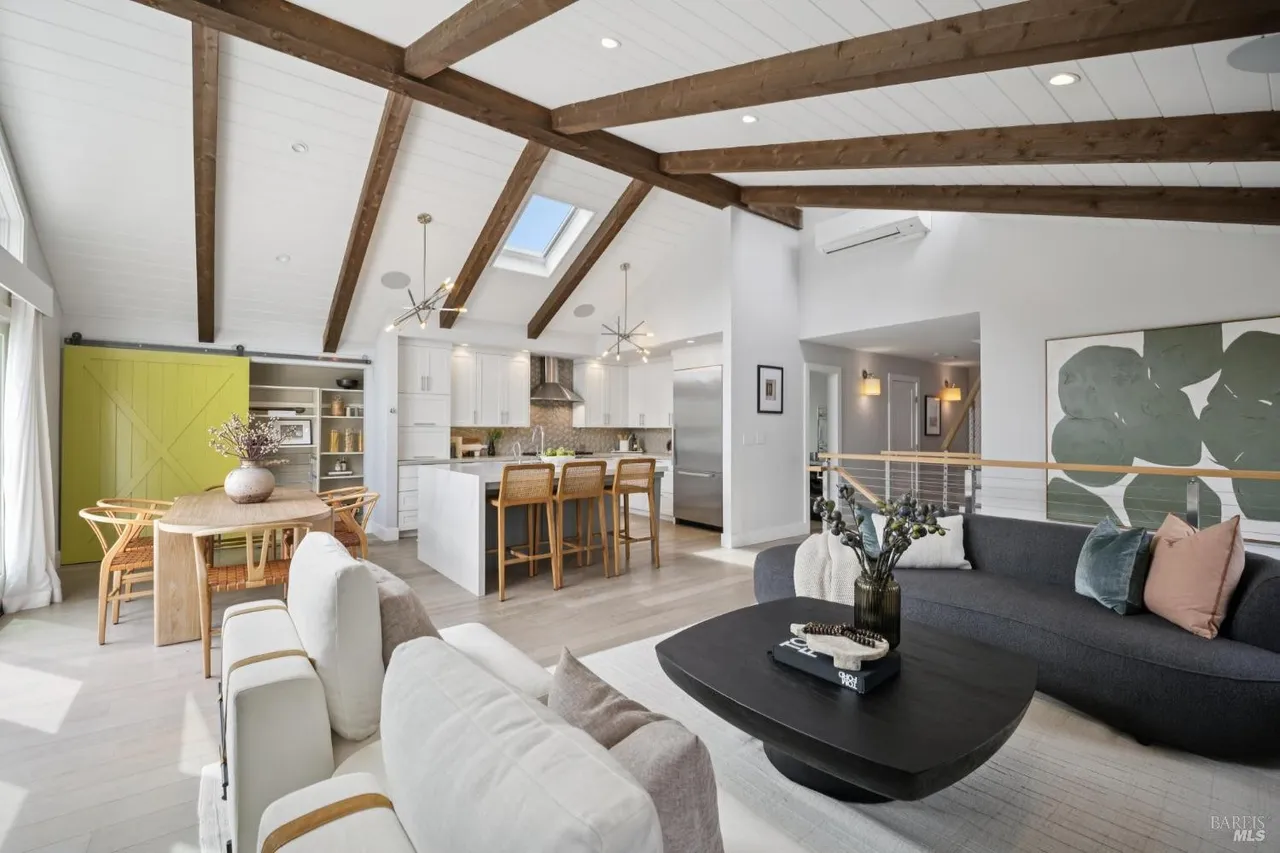 This interior shot showcases an open-concept living space that seamlessly integrates the living room, dining area, and kitchen. The living room features a comfortable gray sofa, a unique dark wood coffee table, and a large abstract painting, while the dining area includes a round wooden table with woven chairs and a striking yellow sliding barn door. The kitchen boasts white cabinetry, a central island with bar stools, and stainless steel appliances, all under a high ceiling with exposed wooden beams and a skylight, creating a bright and airy atmosphere.