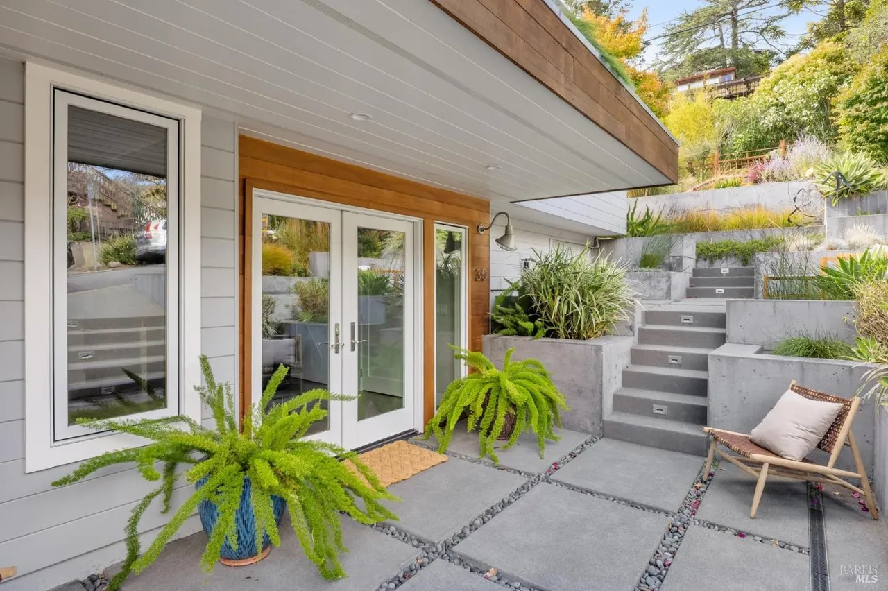 This image showcases the entryway of a modern home, featuring double glass doors framed by wood trim and a large window to the left. Lush green ferns in pots flank the entrance, adding a touch of nature. The walkway is paved with large square stones interspersed with pebbles, leading to concrete steps and tiered garden beds, creating an inviting and stylish approach to the house.