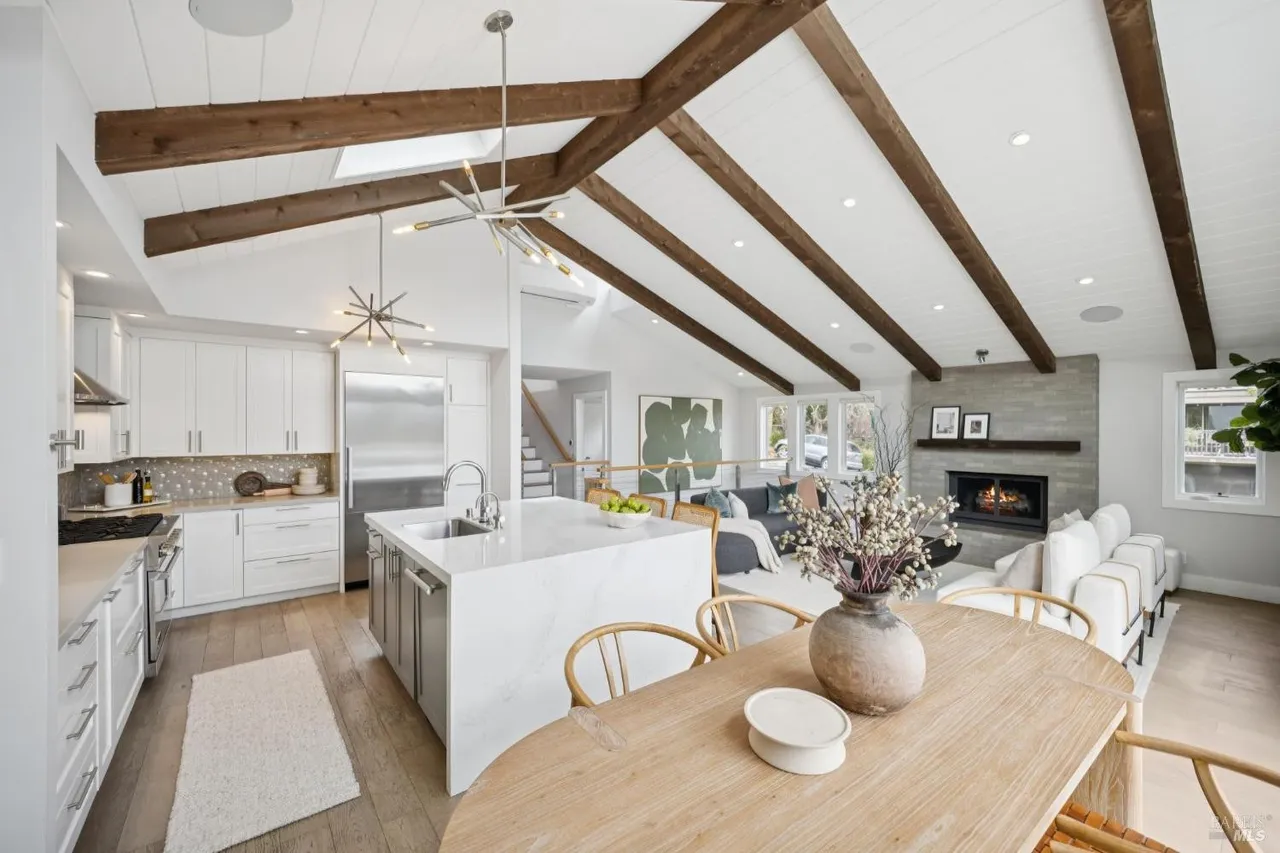This is a bright and airy open-concept kitchen and living space featuring a vaulted ceiling with exposed wooden beams. The kitchen boasts white cabinetry, stainless steel appliances, and a large island with a marble countertop. The living area includes a fireplace with a stone surround and comfortable seating, creating a warm and inviting atmosphere.