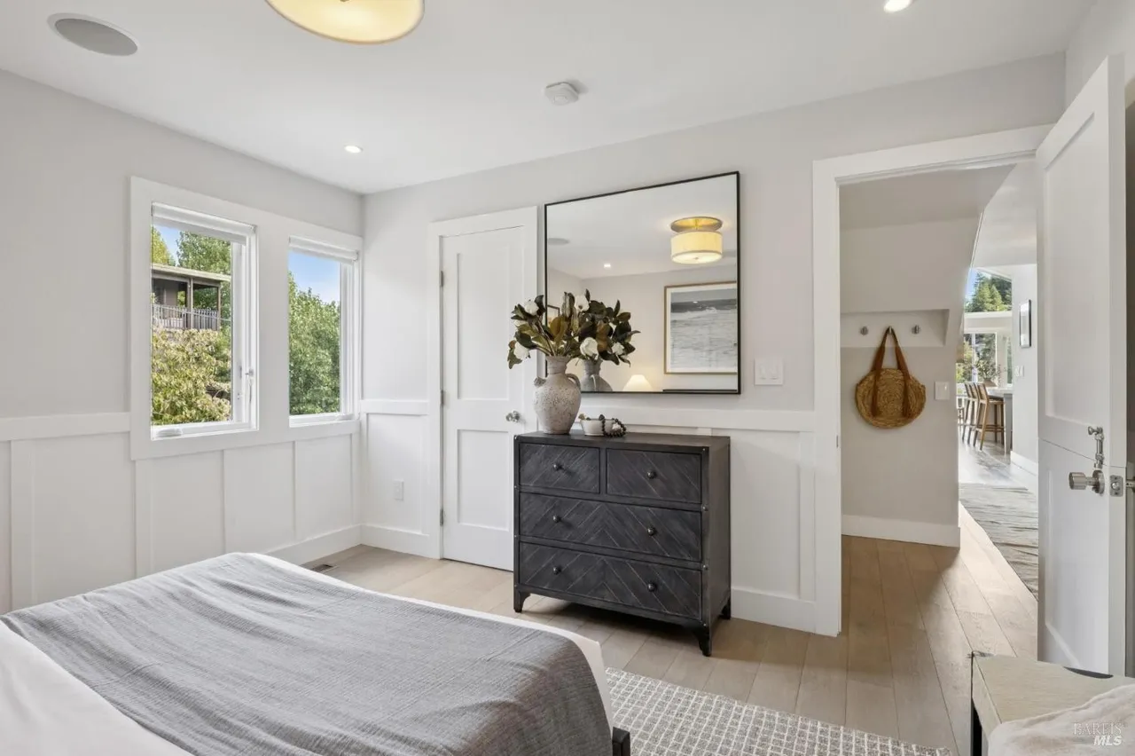 This is a bright and airy primary bedroom featuring white walls with wainscoting, light hardwood floors, and two windows that provide natural light. A dark wood dresser with a mirror above it adds a touch of elegance, while a gray bedspread on the bed creates a cozy and inviting atmosphere. An open doorway leads to another room, enhancing the sense of spaciousness.