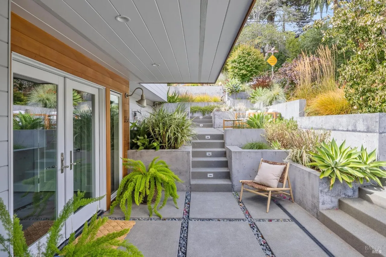 This image showcases a modern patio area with a tiered garden design. The patio features concrete pavers interspersed with decorative stones, leading to a set of concrete stairs with integrated lighting. A stylish chair with a cushion adds a touch of comfort, while lush greenery and tiered concrete planters create a visually appealing and inviting outdoor space.