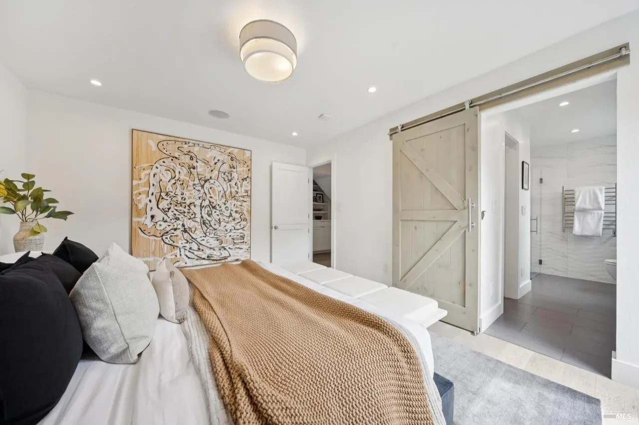 This is a bright and airy primary bedroom featuring a large abstract art piece, a sliding barn door leading to the bathroom, and a comfortable bed with multiple pillows and a cozy throw. The room is painted in a clean white, enhancing the natural light and creating a serene atmosphere. The style is modern and inviting, perfect for relaxation.
