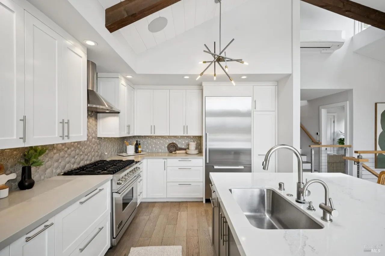 This is a bright and modern kitchen featuring white cabinetry, stainless steel appliances, and light wood flooring. A large island with a stainless steel sink and white countertop anchors the space, while a unique chandelier adds a touch of contemporary style. The kitchen is well-lit and appears spacious, offering a clean and inviting atmosphere.
