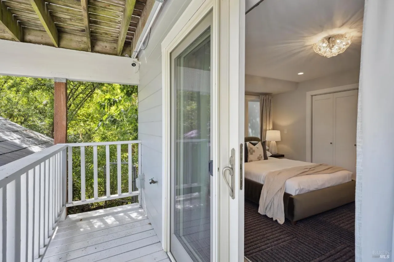 This image showcases a bedroom viewed through a sliding glass door leading to a balcony. The bedroom features a bed with neutral linens, a bedside table with a lamp, and a closet. The balcony offers an outdoor space with white railings and a view of greenery, creating a seamless indoor-outdoor living experience.