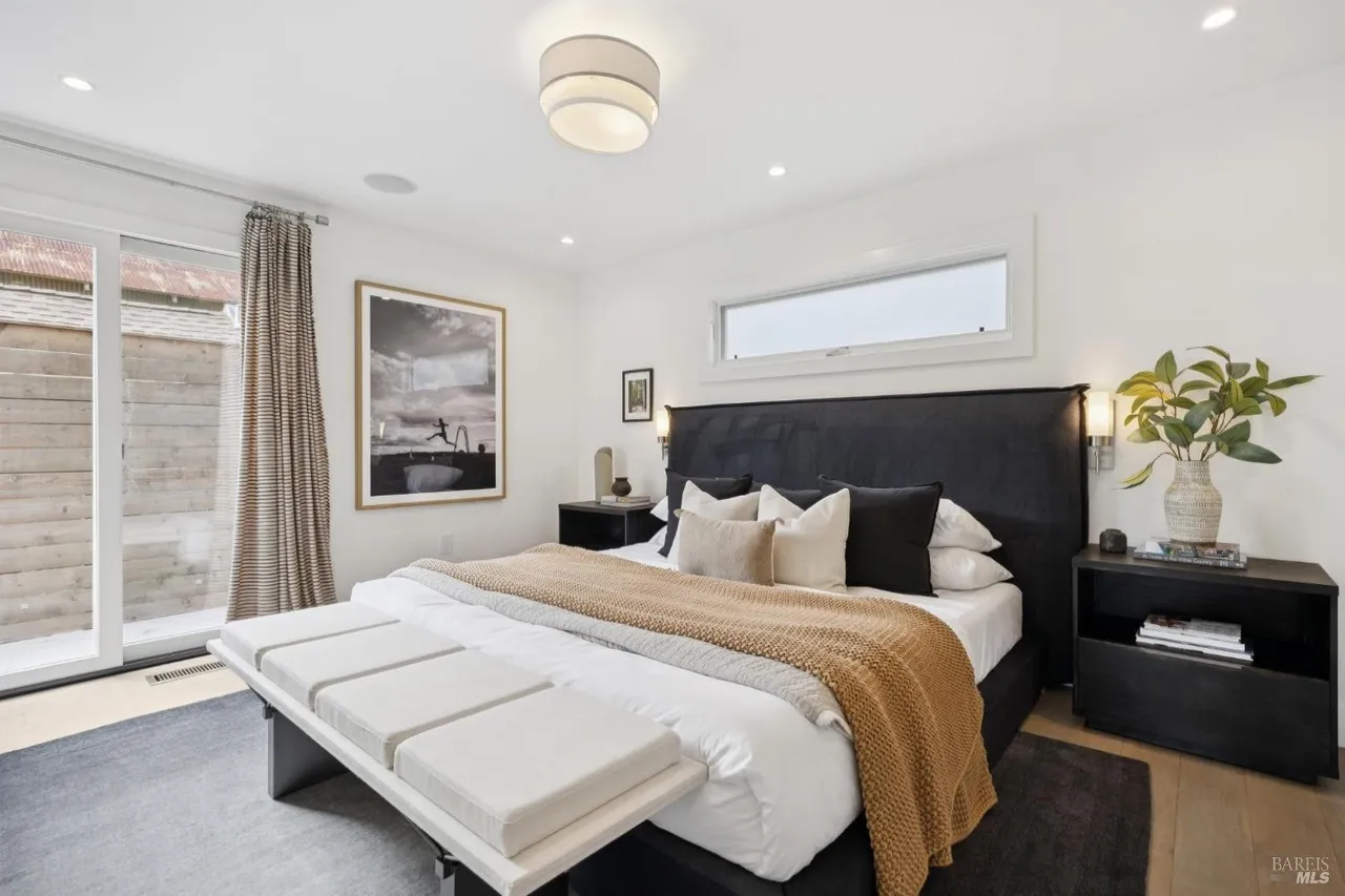 This is a well-lit primary bedroom featuring a large black upholstered headboard, a white duvet with a tan throw, and a bench at the foot of the bed. The room has white walls, a modern ceiling light, and a large framed picture on the wall. There is a sliding glass door to the outside and a small window above the headboard.