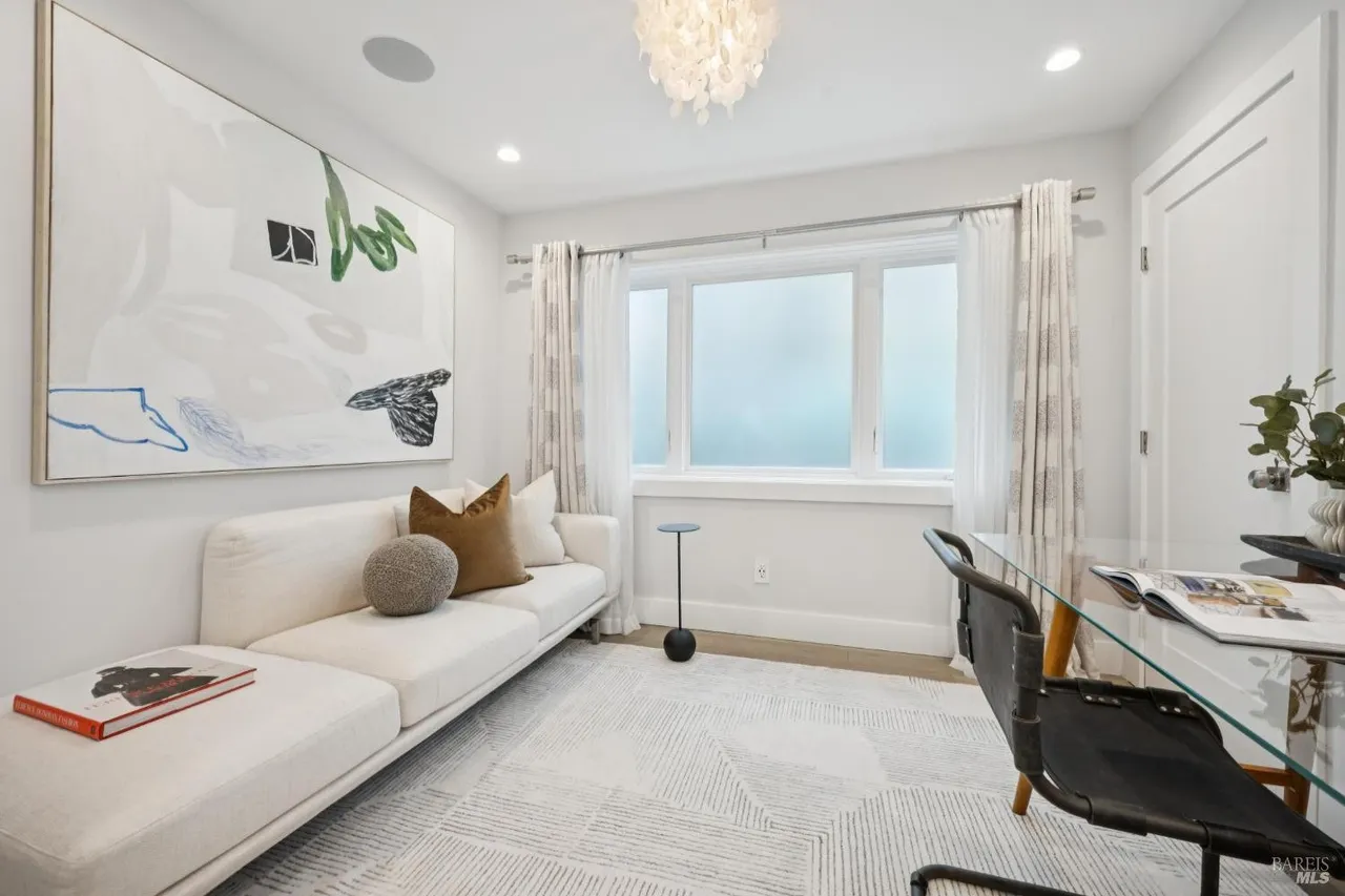 This is an interior shot of a modern office or study. The room features a white sofa with decorative pillows, a large abstract art piece, and a glass-topped desk with a black chair. A patterned rug covers the floor, and natural light filters through a window with sheer curtains, creating a bright and inviting workspace.