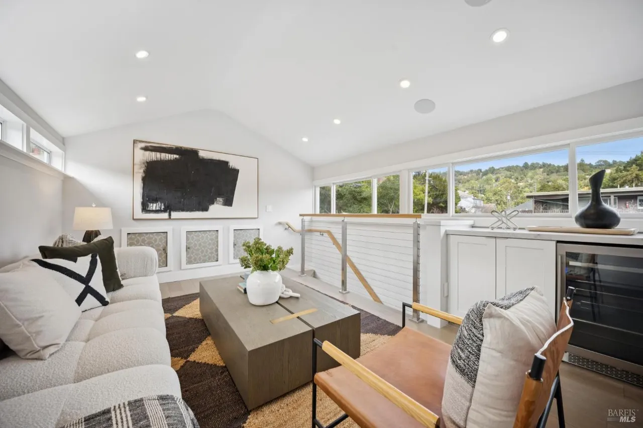 This is an interior shot of a modern living room featuring a white sofa with decorative pillows, a unique geometric coffee table, and a leather chair. A large abstract painting hangs on the wall, complemented by recessed lighting and a view of the outdoors through a series of windows. The room exudes a bright and airy atmosphere with a blend of textures and contemporary design elements.