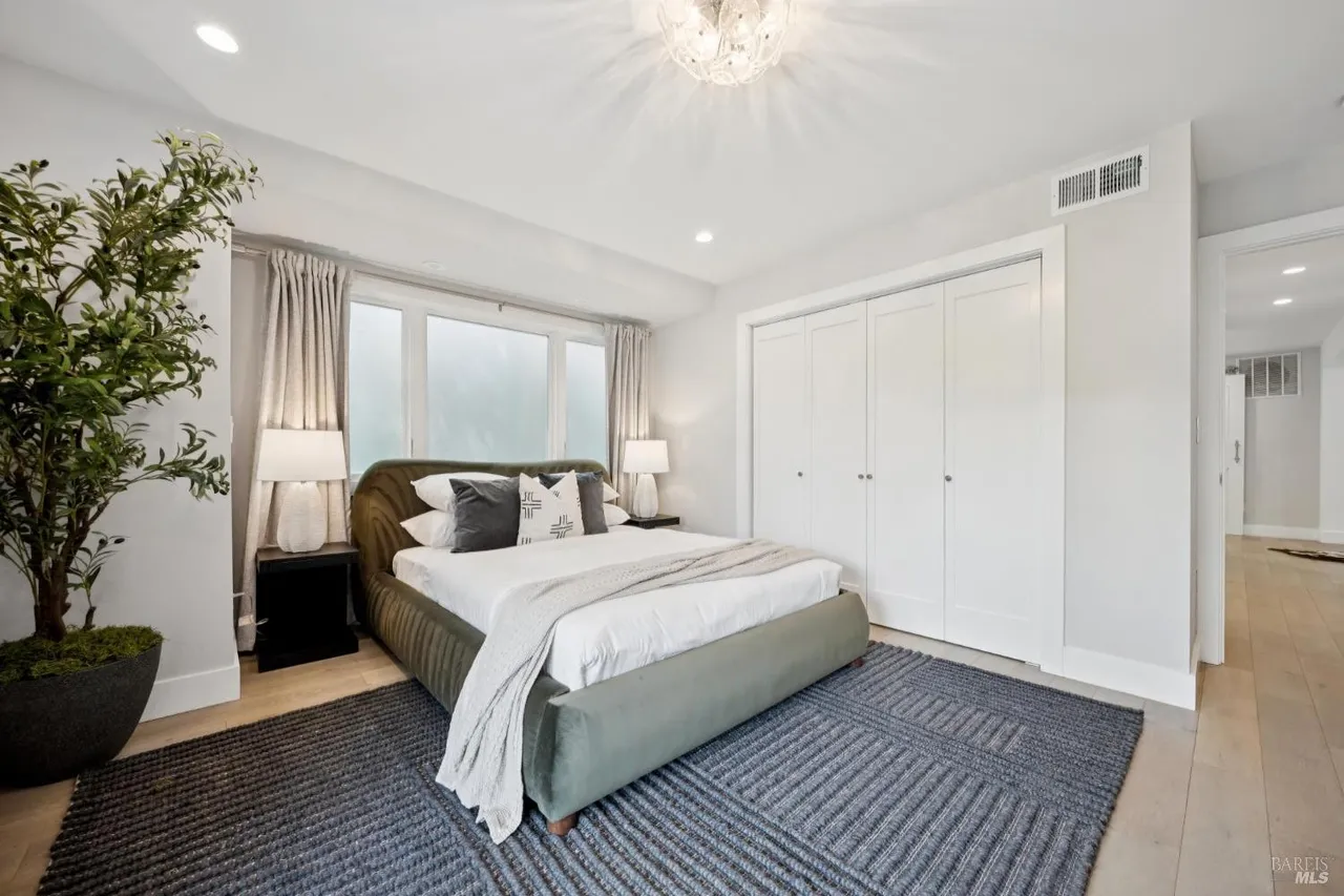 This is a well-lit primary bedroom featuring a green upholstered bed with white linens and decorative pillows, complemented by a gray patterned rug. The room includes built-in closets, a bedside table with a lamp, and a large potted plant, creating a serene and inviting atmosphere. The perspective is from the foot of the bed, showcasing the entire room.