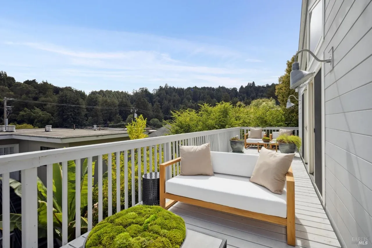 This image showcases a stylish outdoor deck area, featuring a modern wooden-framed sofa with white cushions and neutral-toned pillows. The deck is surrounded by a white railing, offering views of lush greenery and trees in the background. The space is designed for relaxation and outdoor living, enhancing the property's appeal.