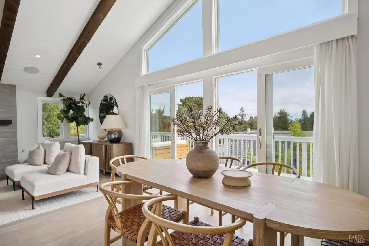 This interior shot showcases a bright and airy dining room with a vaulted ceiling and exposed beams. A large wooden dining table with woven chairs sits in the center, complemented by a decorative vase. Large windows and sliding glass doors provide ample natural light and views of the outdoor scenery, enhancing the room's appeal.
