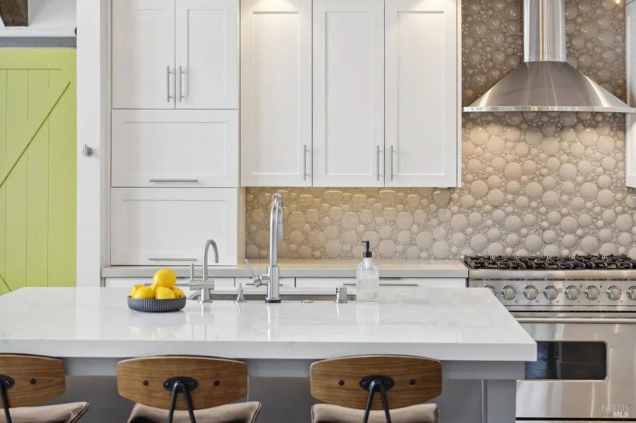 This is a bright and modern kitchen featuring white cabinets, stainless steel appliances, and a large island with a white countertop. The backsplash has a unique circular pattern, and the kitchen includes a gas range with a stainless steel hood. The perspective is from the island looking towards the kitchen appliances.