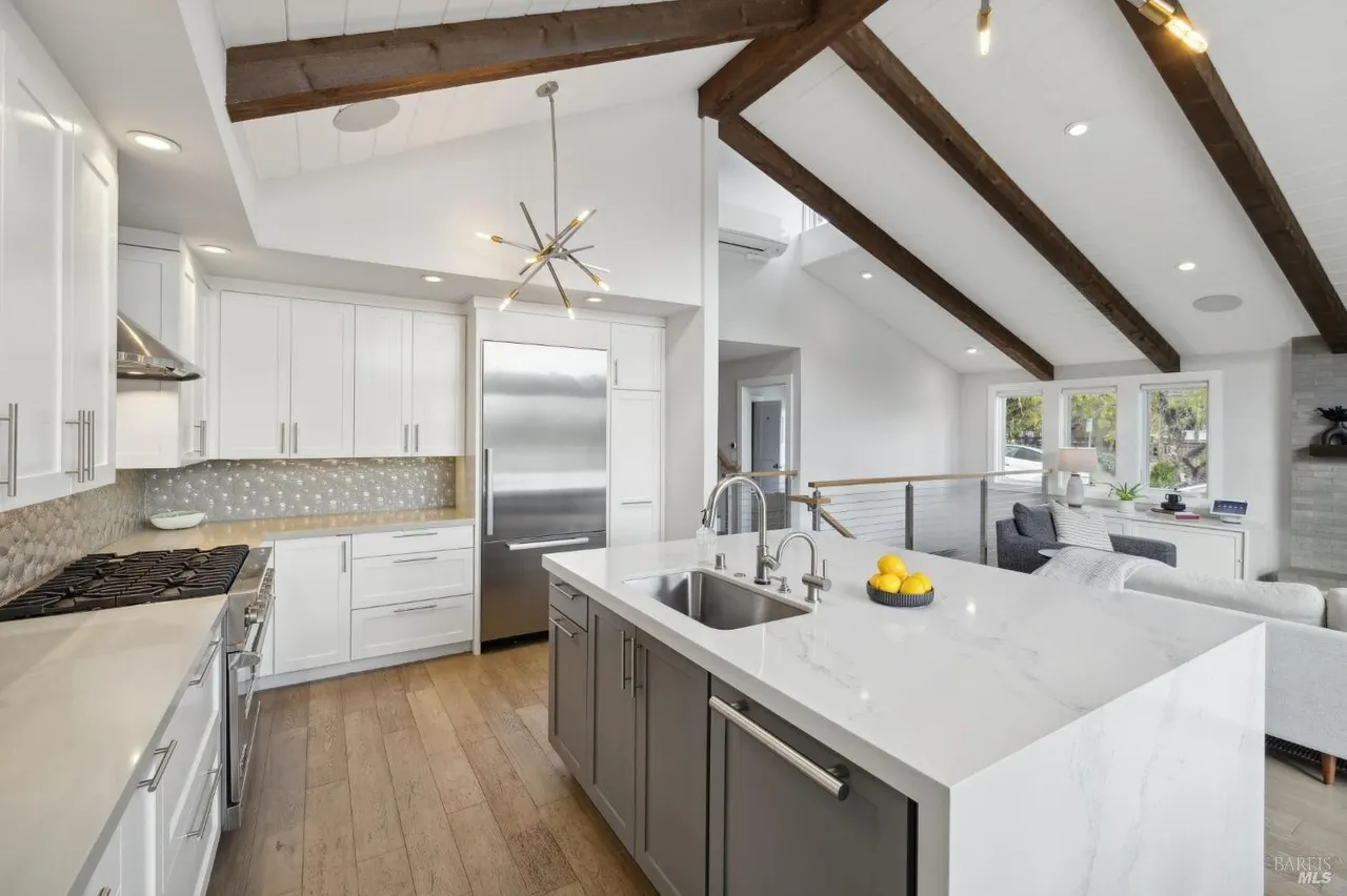 This is a bright and airy kitchen with a modern design. It features white cabinets, stainless steel appliances, a large island with a white countertop, and hardwood floors. The kitchen opens to a living area with exposed wooden beams, creating a spacious and inviting atmosphere.