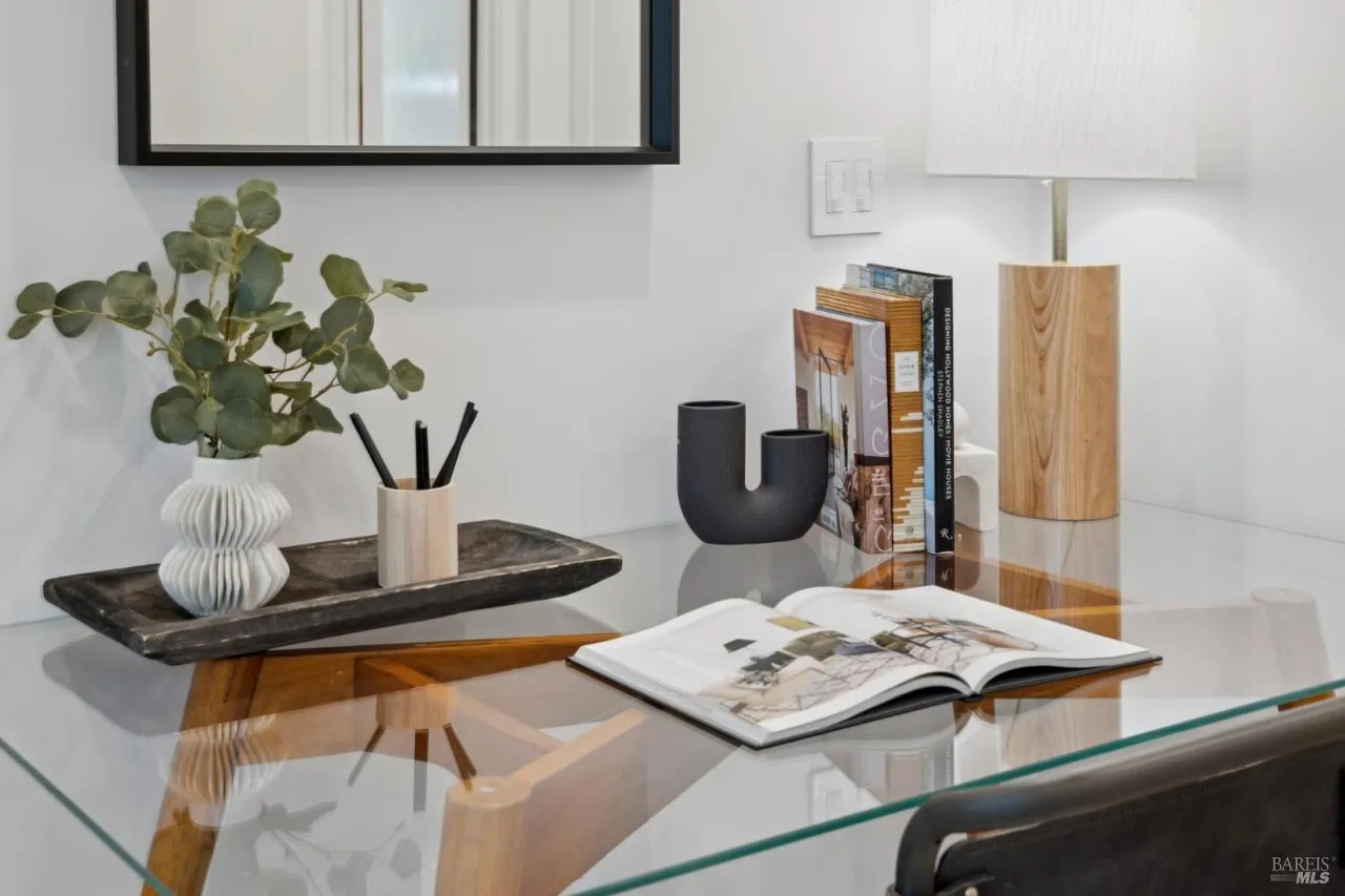 This image showcases a well-organized office space with a glass-topped desk. The desk features decorative elements such as a vase with greenery, a modern black vase, books, and a wooden lamp, creating a stylish and inviting workspace. The composition is clean and bright, emphasizing the functionality and aesthetic appeal of the office.