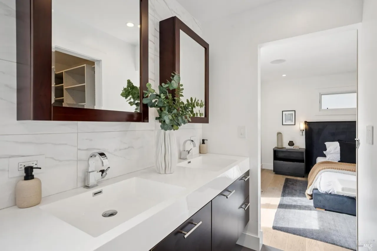 This is a well-lit primary bathroom featuring a double vanity with white countertops and dark wood cabinetry. Two framed mirrors hang above the sinks, and a vase with greenery adds a touch of nature. The bathroom opens into a bedroom, creating a seamless and luxurious feel.