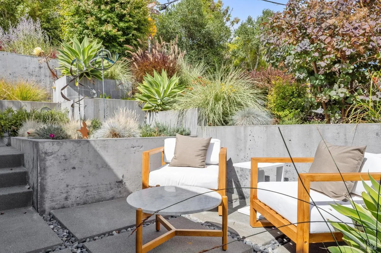 This image showcases a stylish outdoor patio area with modern design elements. Two wooden-framed chairs with white cushions and neutral pillows are arranged around a small round table, creating an inviting seating arrangement. The patio is surrounded by a tiered concrete retaining wall adorned with various plants and greenery, adding a touch of nature and privacy to the space.