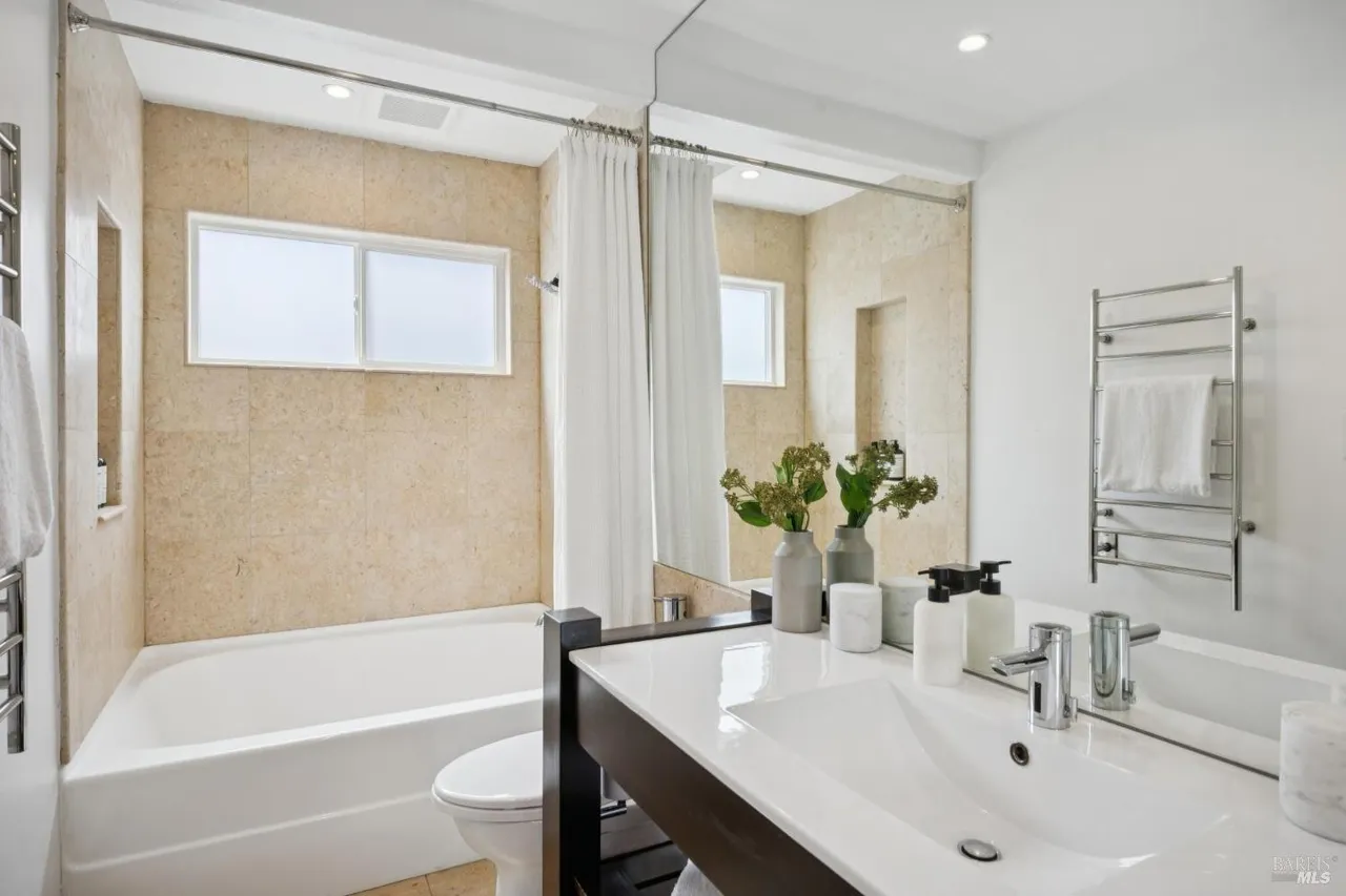 This is a well-lit bathroom featuring a bathtub with a white shower curtain, a window above the tub, and beige tiling on the walls. The vanity has a white countertop with a modern sink and faucet, complemented by decorative vases and toiletries. A heated towel rack is visible on the right, adding a touch of luxury.