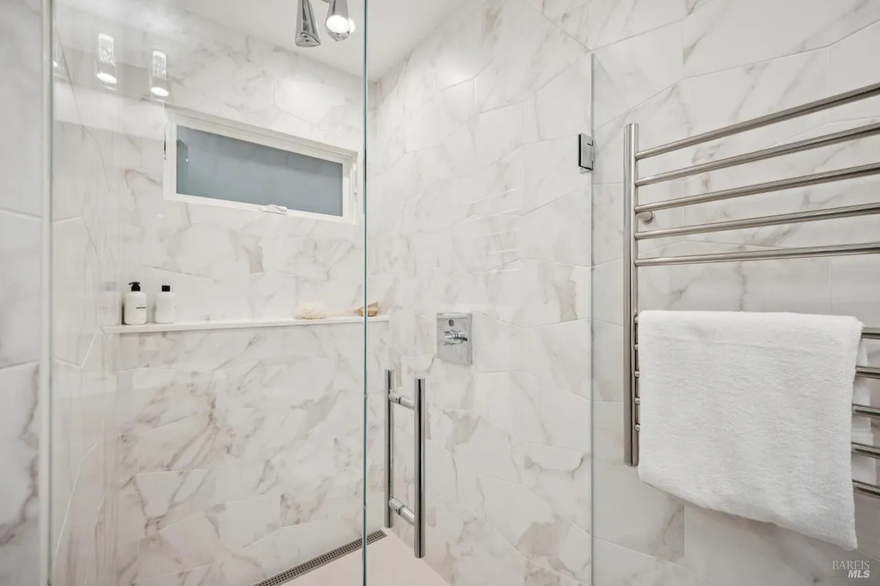 This is a bright and modern primary bathroom featuring marble-patterned walls and a glass-enclosed shower. A sleek, chrome towel warmer holds a fluffy white towel, adding a touch of luxury. The shower includes a built-in shelf with toiletries and a small window for natural light.