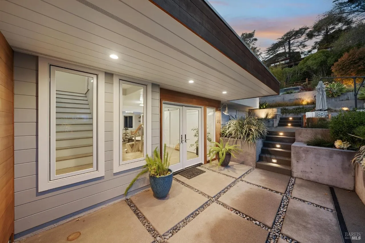 This image showcases the entryway of a modern home, featuring a concrete tile patio with pebble accents, leading to double glass doors. Large windows flank the entrance, offering glimpses of the interior. The exterior is complemented by lush greenery and tiered landscaping, creating an inviting and stylish approach to the residence.