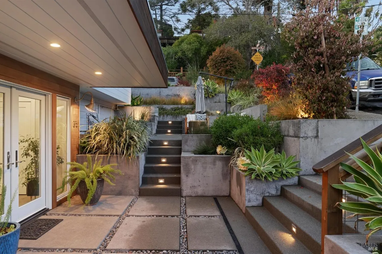 This image showcases an inviting entryway with modern landscaping. Concrete steps with embedded lighting lead up to the property, flanked by raised garden beds filled with lush greenery. The pathway is paved with concrete squares interspersed with gravel, creating a visually appealing and functional space.