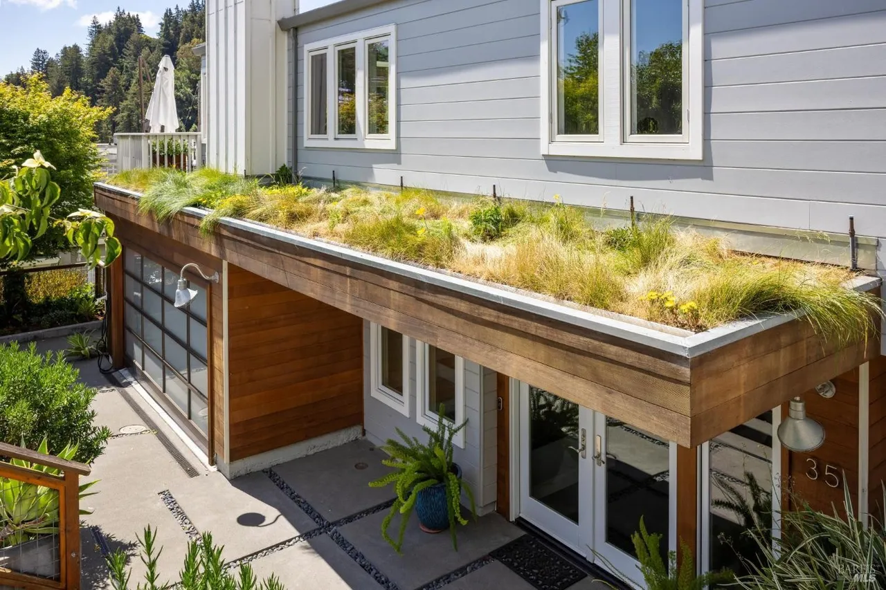 The image showcases the entryway of a modern home, featuring a unique green roof over the entrance. The exterior is clad in light gray siding complemented by wood accents around the garage and entryway. A paved walkway leads to the double doors, enhancing the property's curb appeal.