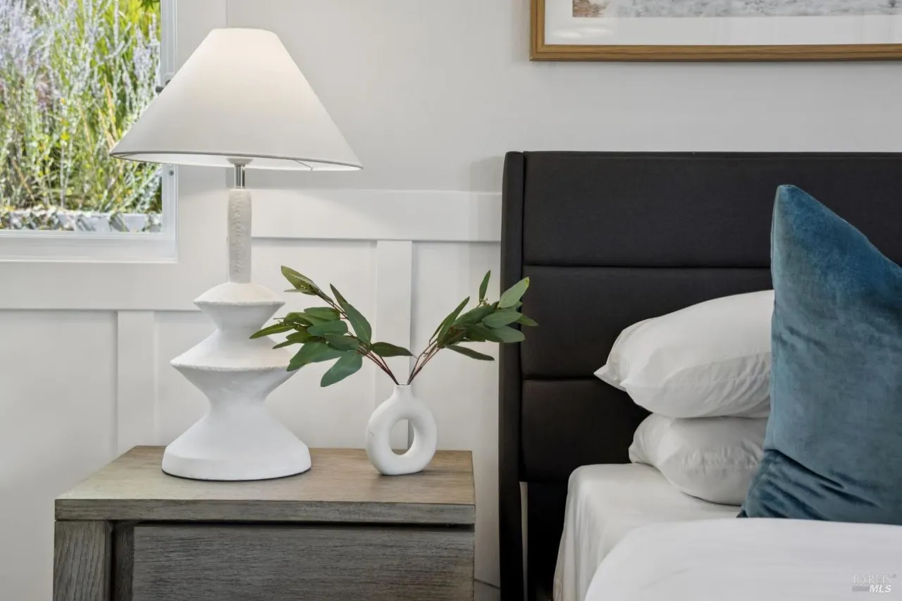 This image showcases a well-staged bedroom featuring a dark upholstered headboard, crisp white linens, and a decorative blue pillow. A bedside table holds a modern white lamp and a small vase with greenery, adding a touch of nature. The room exudes a clean and inviting atmosphere, perfect for a guest bedroom.