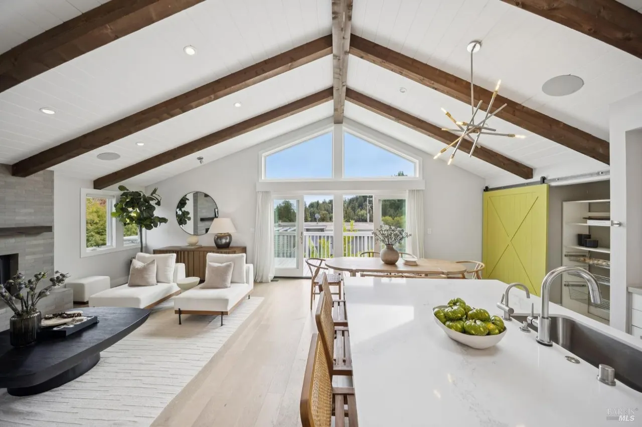 This is an interior shot of a modern living room and kitchen area featuring a vaulted ceiling with exposed wooden beams. The living room area includes white chaise lounges, a dark coffee table, and a light-colored rug, while the kitchen area has a white countertop island with bar stools and a view of the dining area. The overall impression is bright, airy, and stylish.