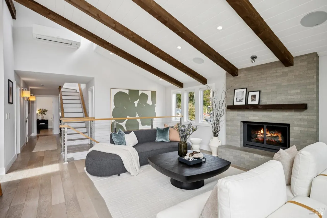 This is an interior shot of a living room featuring a fireplace, a gray sofa, and a white sofa. The room has wooden beams on the ceiling and a large piece of artwork on the wall. The overall style is modern and inviting, with a focus on natural light and comfortable furnishings.