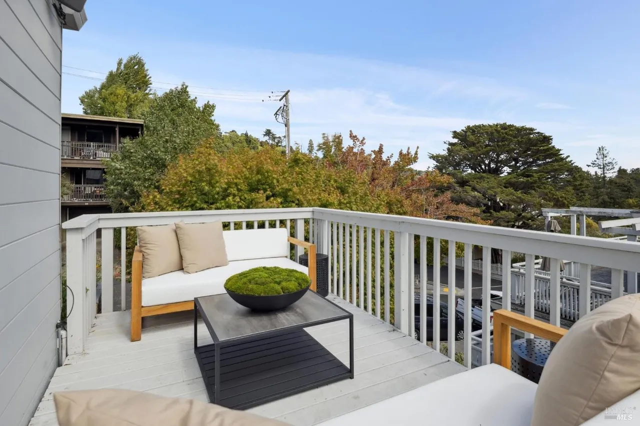 This image showcases a stylish outdoor balcony or deck area. The space is furnished with modern white seating accented by wooden frames and neutral-toned pillows. A sleek, dark-colored coffee table with a decorative moss centerpiece adds a touch of elegance. The white railing provides a clean, bright border, framing a view of lush greenery and a clear blue sky, creating an inviting and relaxing atmosphere.
