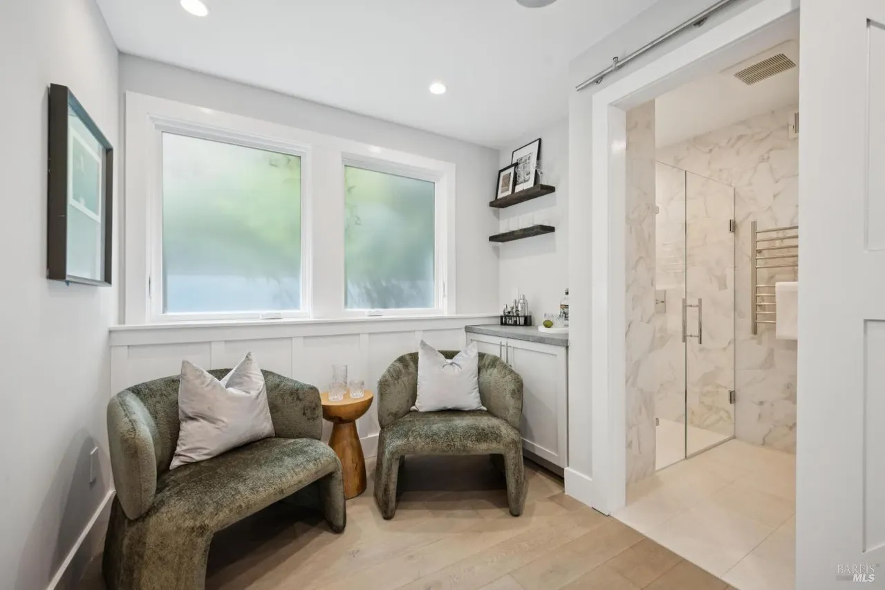 This is a bright and stylish guest bathroom featuring two comfortable armchairs with pillows, a small wooden side table, and a built-in bar area. The bathroom leads into a modern shower with glass doors and marble-like tiling. The room is well-lit with natural light from the frosted windows, creating a relaxing and inviting atmosphere.