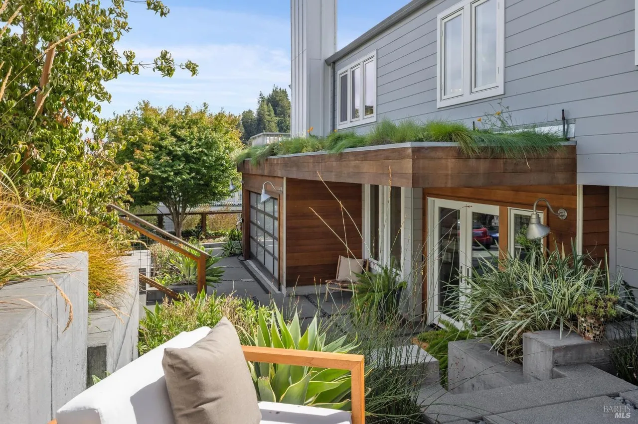 This image showcases a beautifully landscaped yard and garden area adjacent to a modern home. The exterior features a combination of wood and gray siding, with a green roof element adding a unique touch. The patio area is furnished with stylish outdoor seating, creating an inviting and relaxing atmosphere.