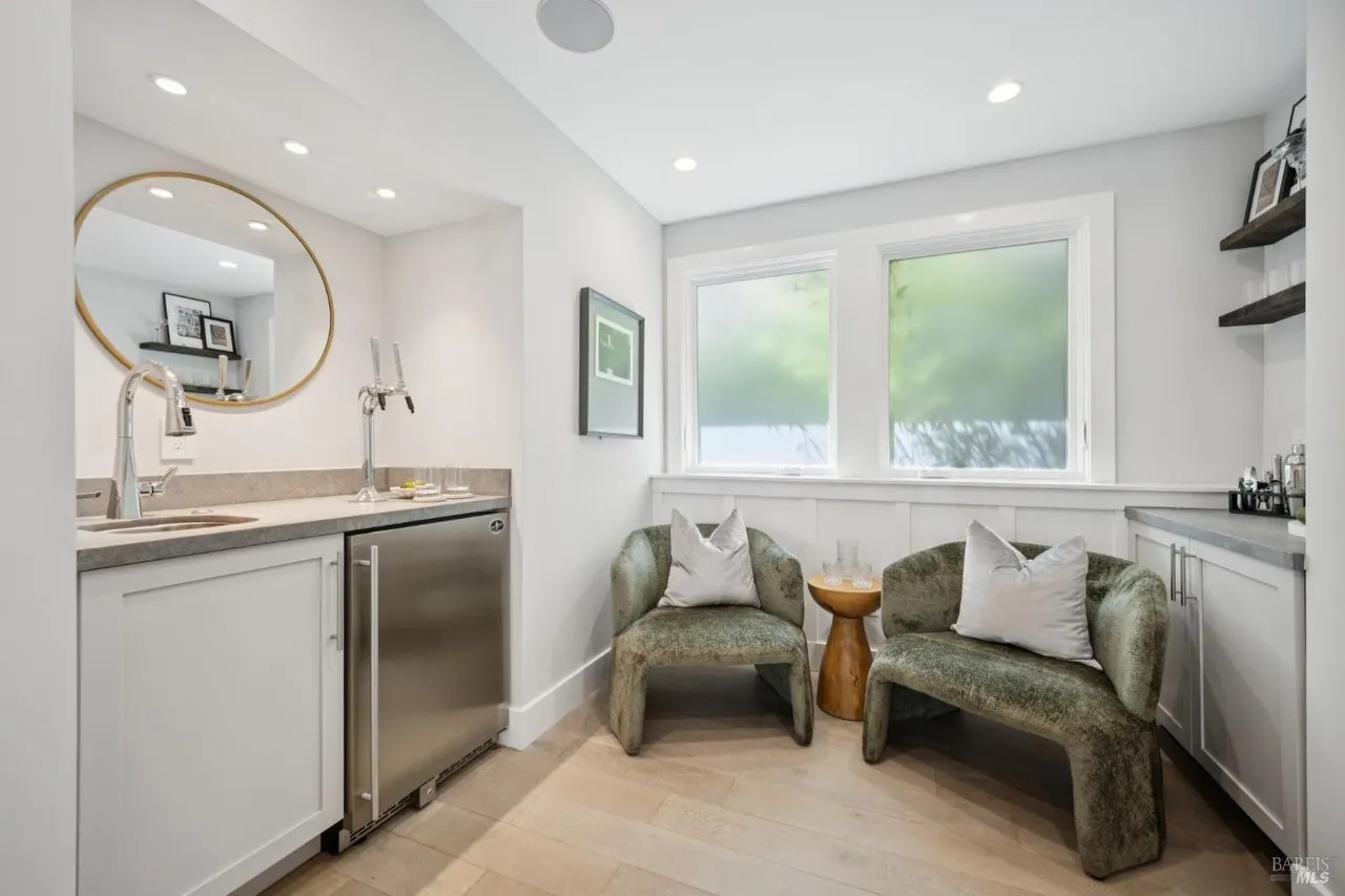 This interior shot showcases a stylish wine cellar or bar area, featuring a sink, beverage refrigerator, and custom cabinetry. Two comfortable chairs flank a small side table, creating an inviting space for relaxation and entertainment. The room is well-lit with recessed lighting and natural light from the windows, enhancing the overall ambiance.