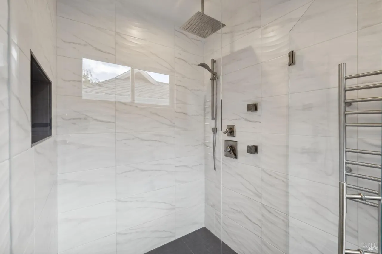 This is a modern primary bathroom featuring a walk-in shower with white marble-style tiling and dark gray flooring. The shower includes a rainfall showerhead, handheld sprayer, and built-in niche for storage. A glass partition separates the shower area, and a heated towel rack is visible on the right.