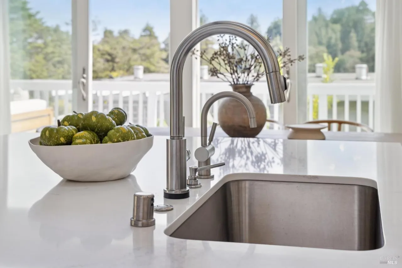 This is a detailed shot of a kitchen sink area, featuring a stainless steel sink and faucet set against a bright white countertop. A bowl of green gourds adds a touch of color, while a vase with dried flowers sits in the background, enhancing the homey feel. The large window behind the sink provides natural light and a view of the outdoors.