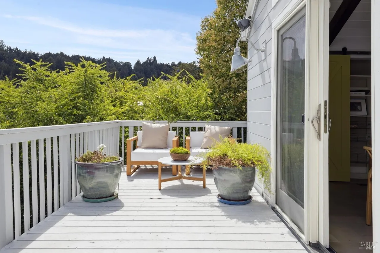 This image showcases a charming outdoor deck area, furnished with two cushioned chairs and a small round table, creating an inviting space for relaxation. Potted plants add a touch of greenery, while the white railing and deck flooring contribute to a clean and airy aesthetic. The backdrop features lush trees, enhancing the sense of privacy and tranquility.