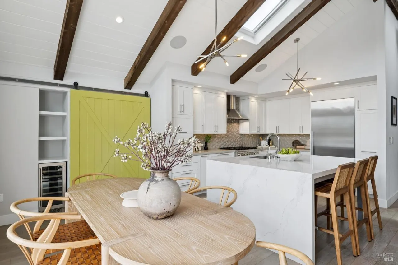 This is a bright and airy kitchen featuring white cabinets, stainless steel appliances, and a large island with a marble countertop. The kitchen also has wooden beams on the ceiling and a unique light fixture. A wooden dining table with chairs is in the foreground, adding a cozy touch to the modern design.