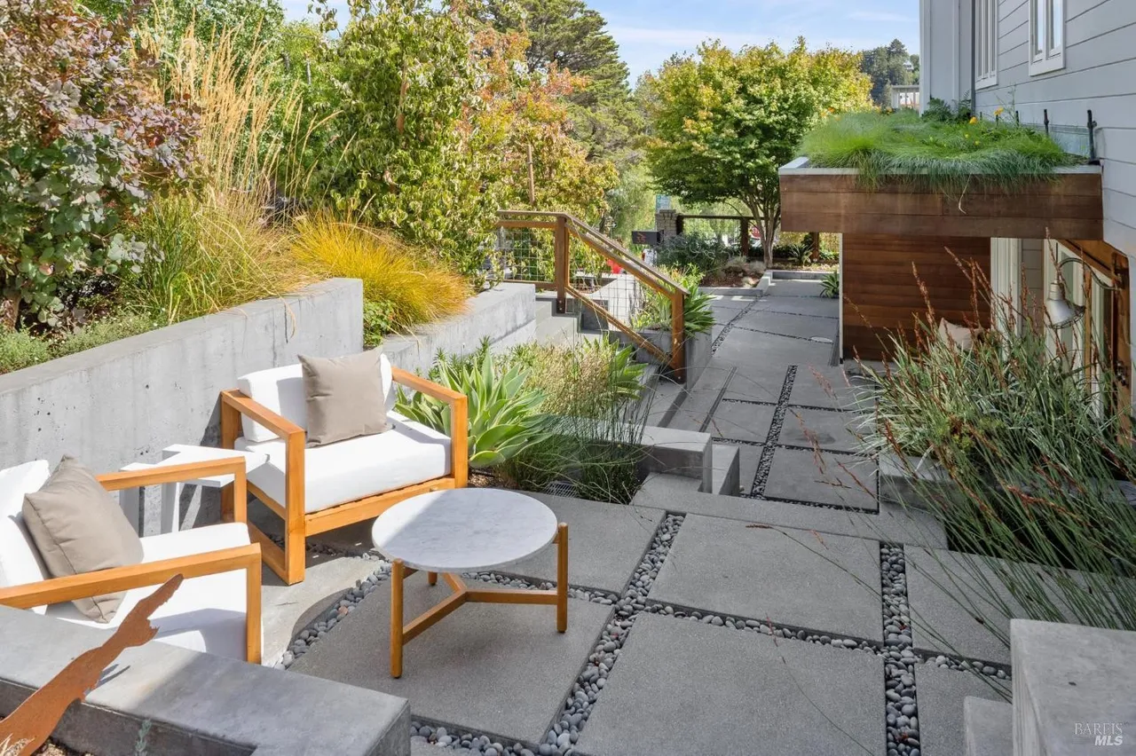 This image showcases a beautifully designed outdoor patio area. The space features modern concrete pavers interspersed with decorative stones, creating a visually appealing and low-maintenance surface. Comfortable outdoor seating is arranged around a small table, suggesting a relaxing and inviting atmosphere for entertaining or enjoying quiet moments outdoors.