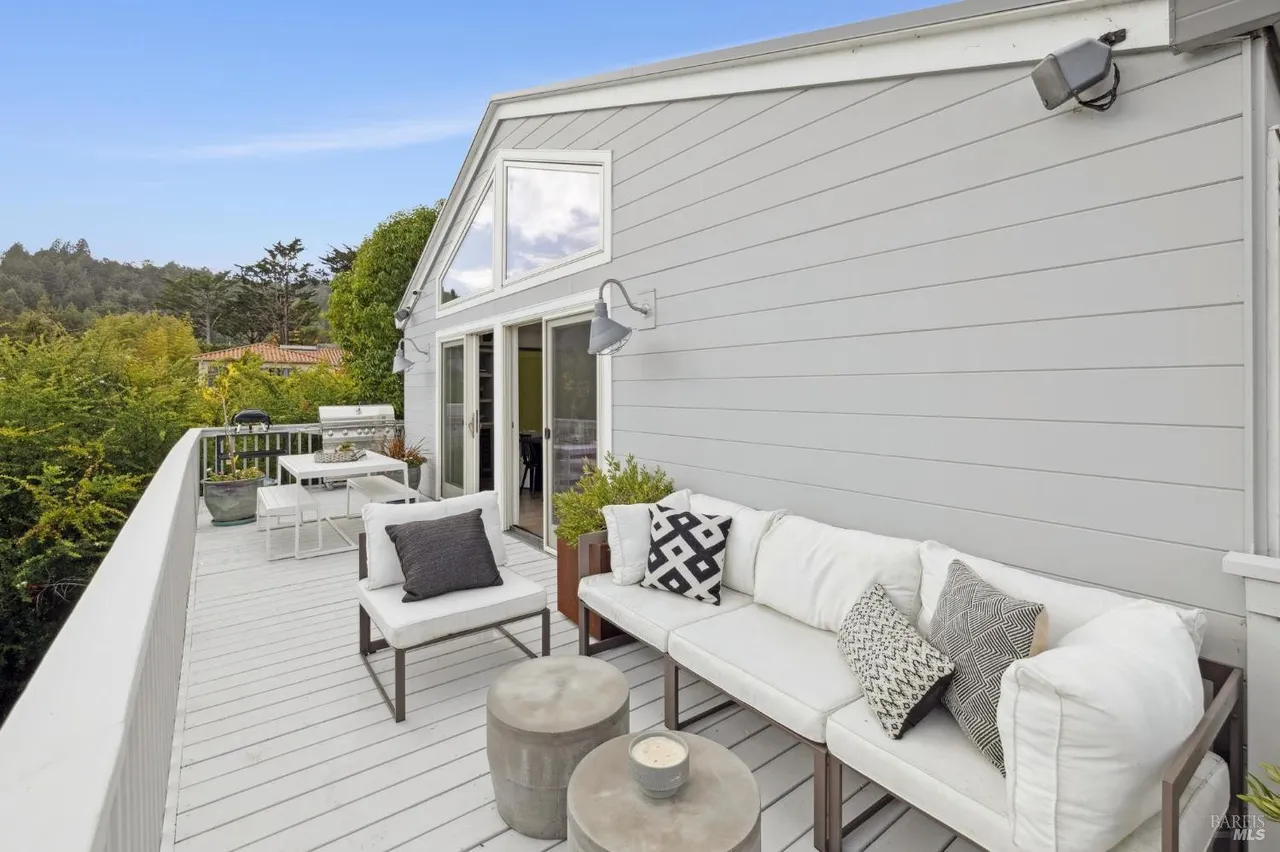 This image showcases a stylish outdoor deck area, featuring a comfortable white sofa with decorative pillows, a modern chair, and concrete side tables. The deck is painted white and overlooks a lush green landscape. The exterior of the house is visible, with gray siding and large windows, creating a serene and inviting outdoor living space.