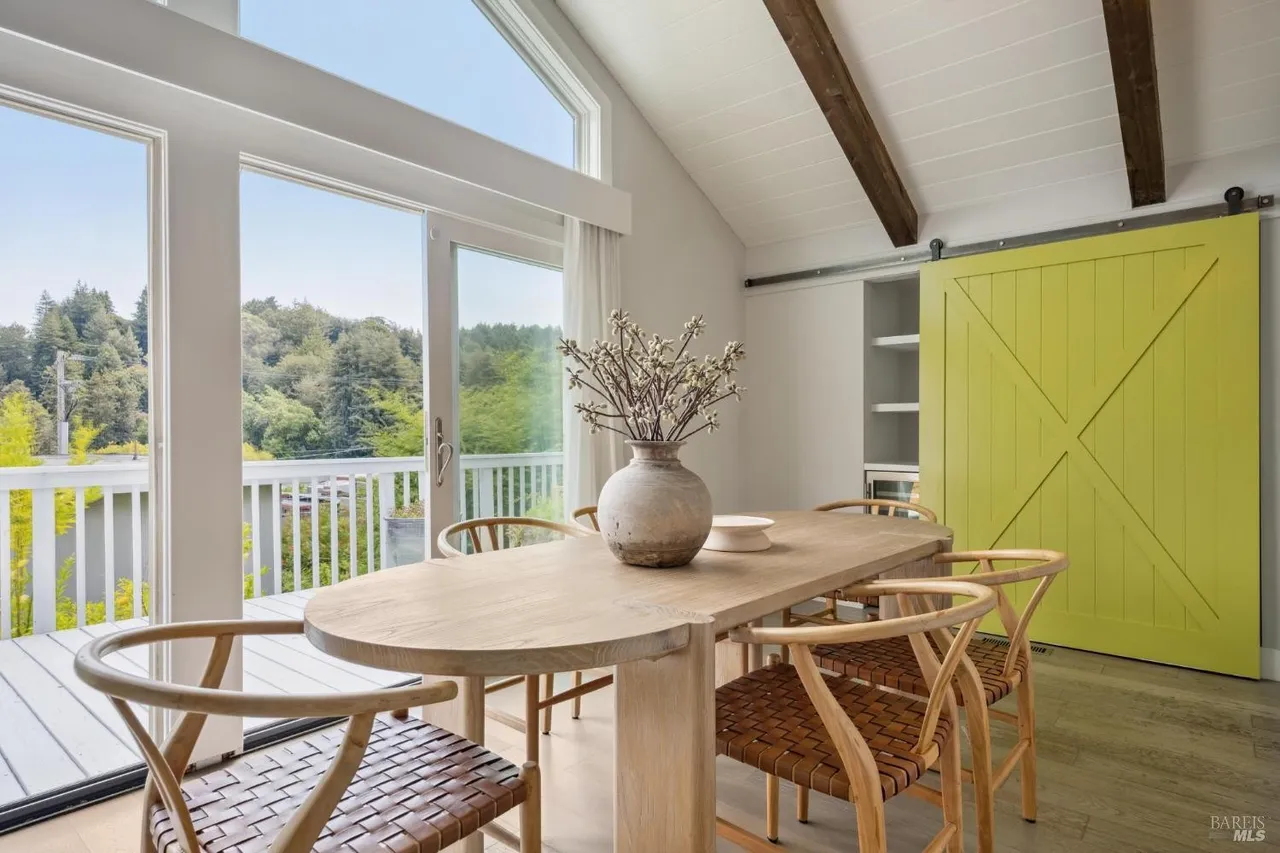 This is an interior shot of a dining room featuring a large, oval wooden table surrounded by woven-seat chairs. A large window offers a view of the outdoors, and a bright yellow barn door adds a pop of color to the room. The room has a modern, rustic feel with exposed beams on the ceiling.