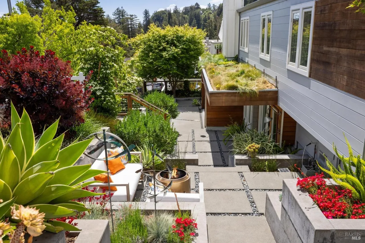 This image showcases a beautifully landscaped yard and garden area. A modern outdoor seating area with a fire pit is visible, surrounded by lush greenery, including various plants and trees. The design incorporates concrete pavers and gravel pathways, leading towards a building with a green roof, creating a serene and inviting outdoor space.