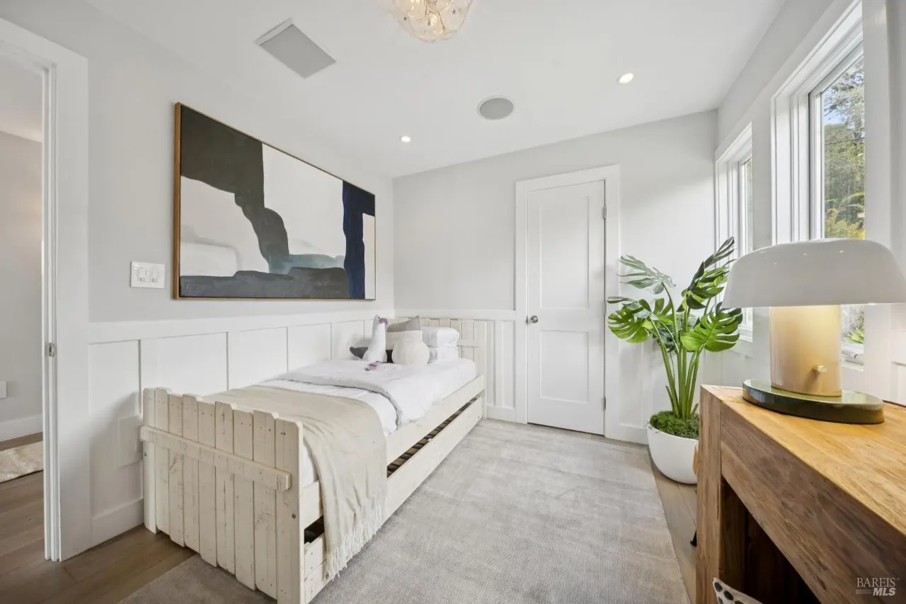 This is a bright and airy guest bedroom featuring a single bed with white bedding and a neutral-toned throw. A large abstract painting hangs above the bed, adding a touch of modern art. The room is well-lit with natural light from the window and features a potted plant, enhancing the room's fresh and inviting atmosphere.