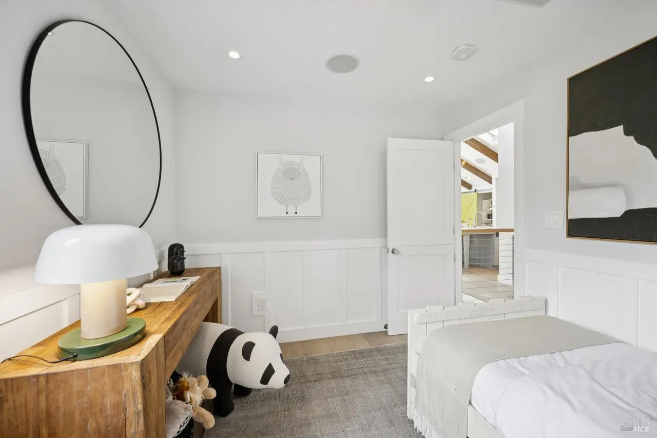 This is a bright and cozy bedroom featuring a single bed with white linens and a gray throw. A wooden desk with a white lamp sits against the wall, accompanied by a round mirror and a stuffed panda. The room has white walls with wainscoting and a gray rug, creating a clean and inviting space.